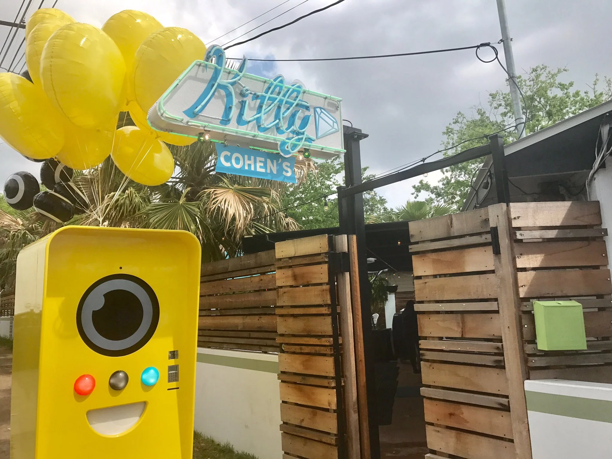 Yellow Snapbot vending machine with balloons beside the neon Kitty Cohen's sign at a patio bar entrance